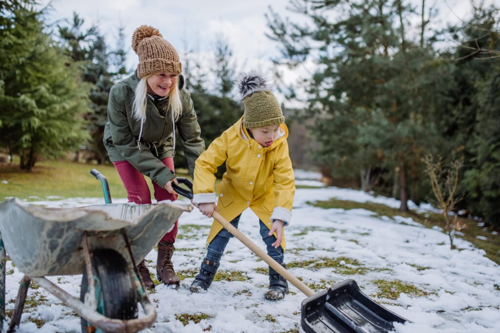 A woman and a child using a shovel and wheelbarrow to clear a lawn with snow on the ground.