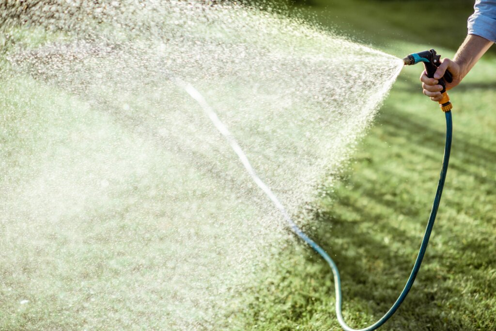 A hand holding a hose nozzle spraying water in a wide arc over a green lawn, emphasizing the importance of knowing when to water after fertilizing.