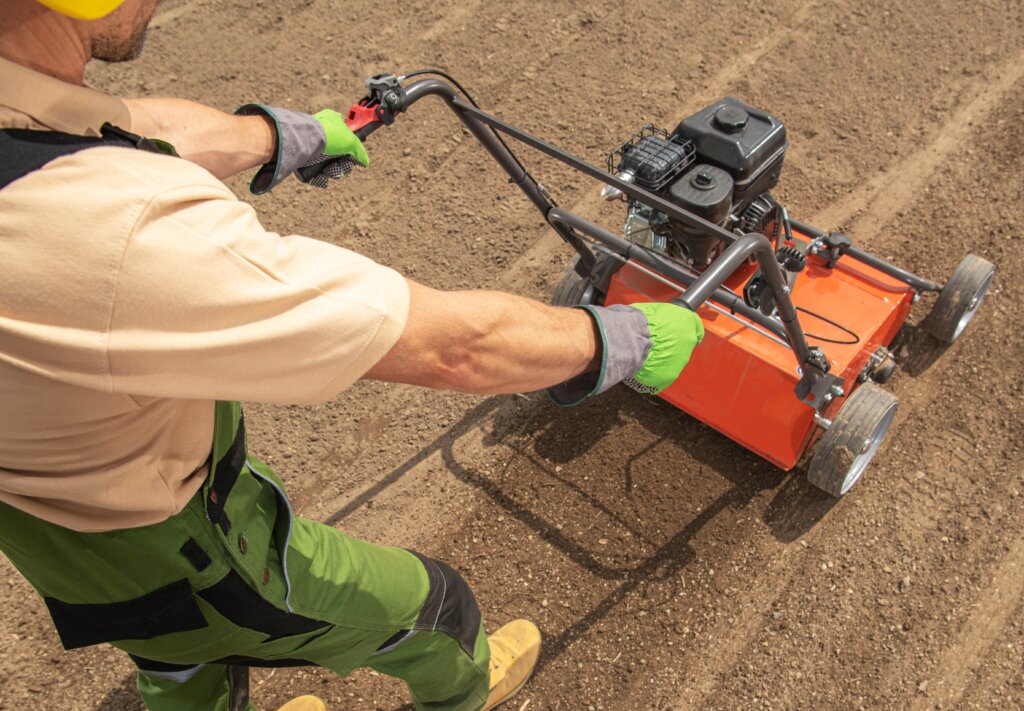 A side-view of a person operating a heavy, motorized core aerator on dry, tilled soil with a residential house in the distance, indicating the intensive effort involved in annual aeration for the question of how often should you aerate your lawn.