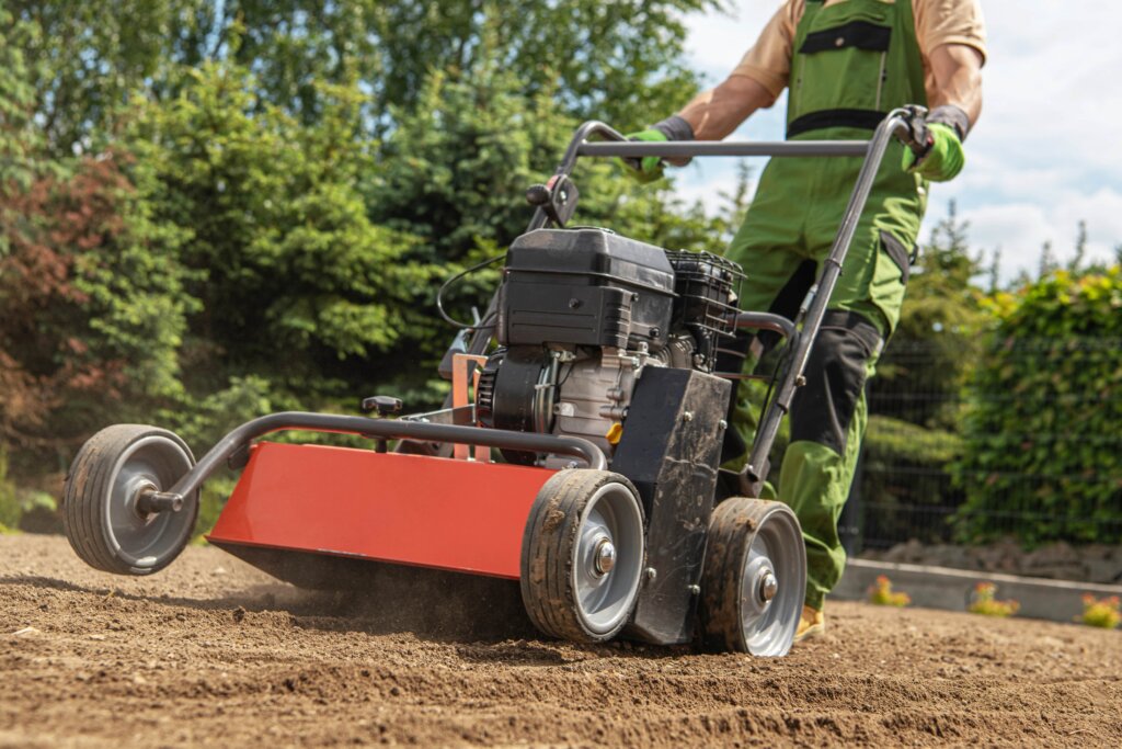 lawn professional in green work gear pushing a heavy, motor-driven core aerator across a patch of bare, prepared soil, highlighting a crucial tool used as a lawn aerator.