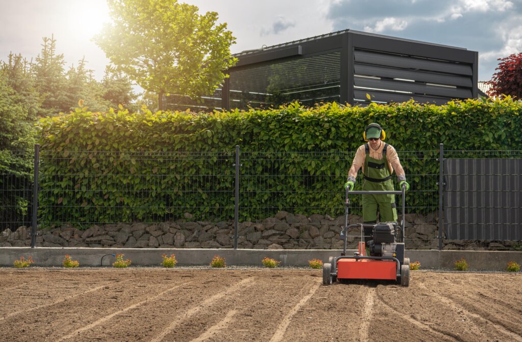 A lawn care professional operates a large, red core aerator machine on a large field of bare dirt, illustrating the intensive process and the question of when should you aerate your lawn.