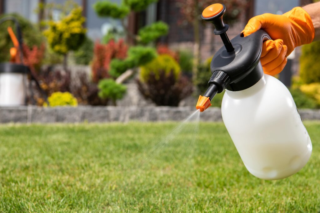 A hand wearing an orange glove holds a small white pump sprayer over a green lawn and landscaped area, demonstrating a common tool for liquid fast release fertilizer.
