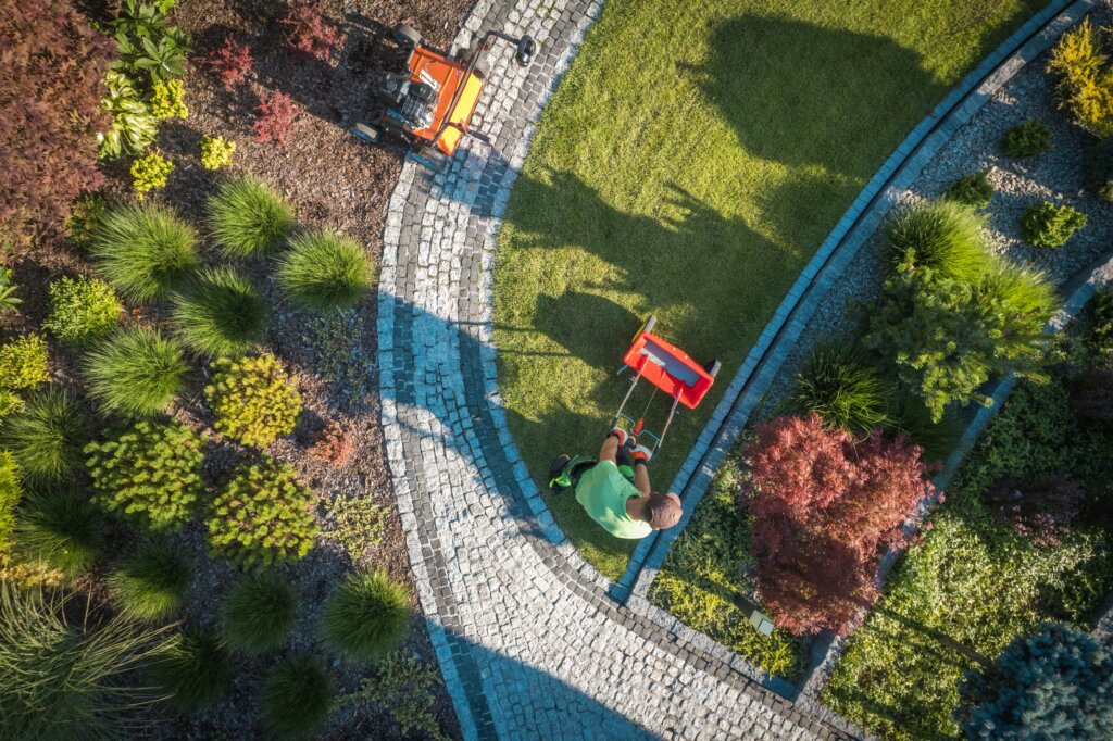 An aerial view of a person pushing a red fertilizer spreader on a green lawn with a curved cobblestone path and landscaped garden beds, illustrating planned fertilizer application.