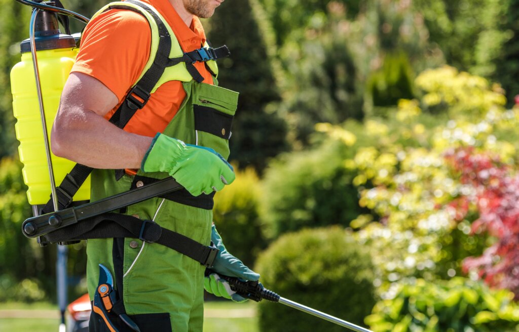 A person wearing green overalls, gloves, and a backpack sprayer is applying liquid to a garden, showcasing one of the effective methods of fertilizer application.