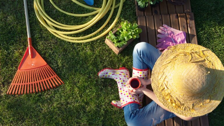 An overhead image of a person sitting on a garden bench, enjoying a cup of coffee. Nearby are pink garden gloves, a variety of plants, a coiled garden hose, and a red garden rake, all representing the tools and results of diligent yard maintenance