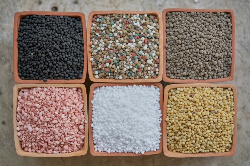 Six terracotta bowls, seen from above, filled with granular fertilizers of varying colors, including black, white, pink, and yellow pellets, representing different types of fertilizers.