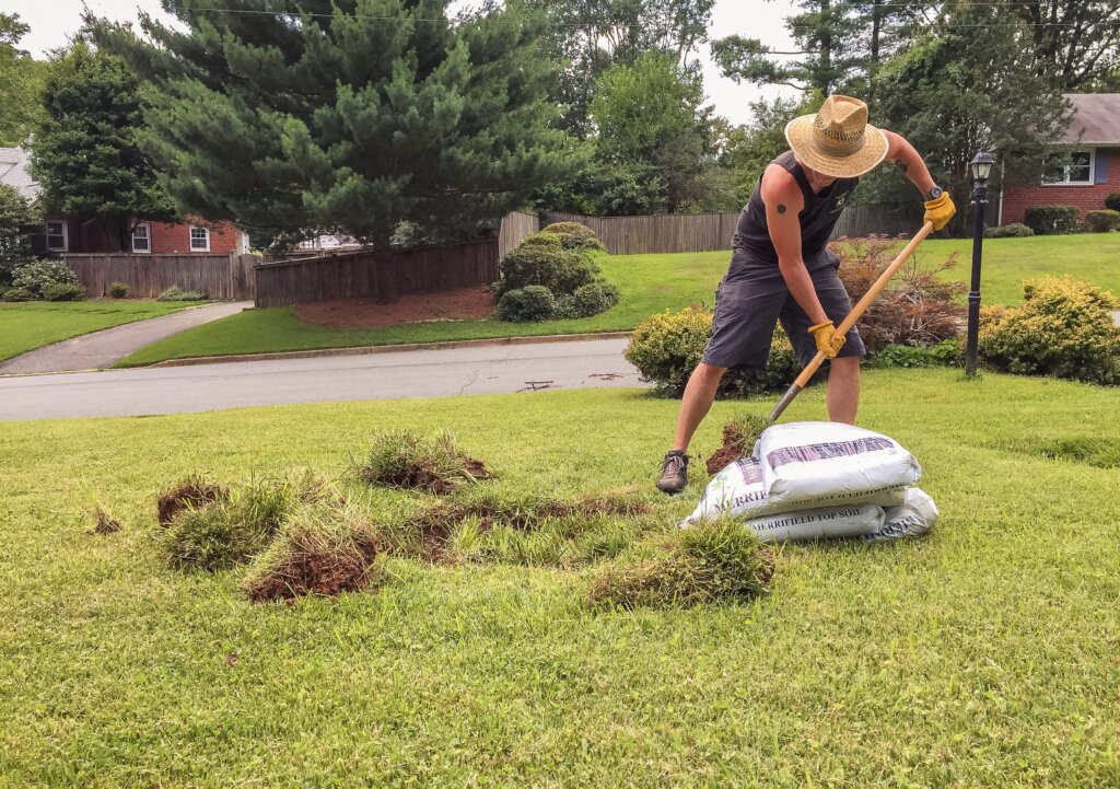 A man wearing a straw hat and gloves uses a shovel to cut away old grass next to a pile of bagged sod, representing the preparation for using fertilizer spikes in a new or renovated area.