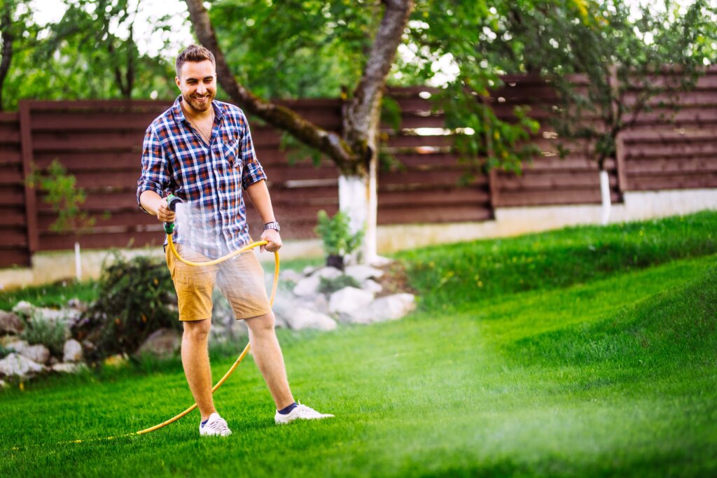A smiling man in a plaid shirt and shorts is watering a green lawn with a yellow hose, demonstrating a key step in how to apply lawn fertilizer.
