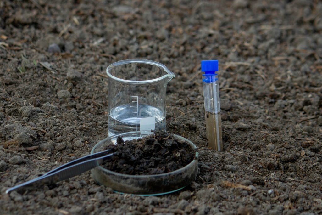 Laboratory glassware including a beaker of water and a test tube of soil sample sitting on dark dirt, symbolizing the first step in learning how to apply lawn fertilizer.