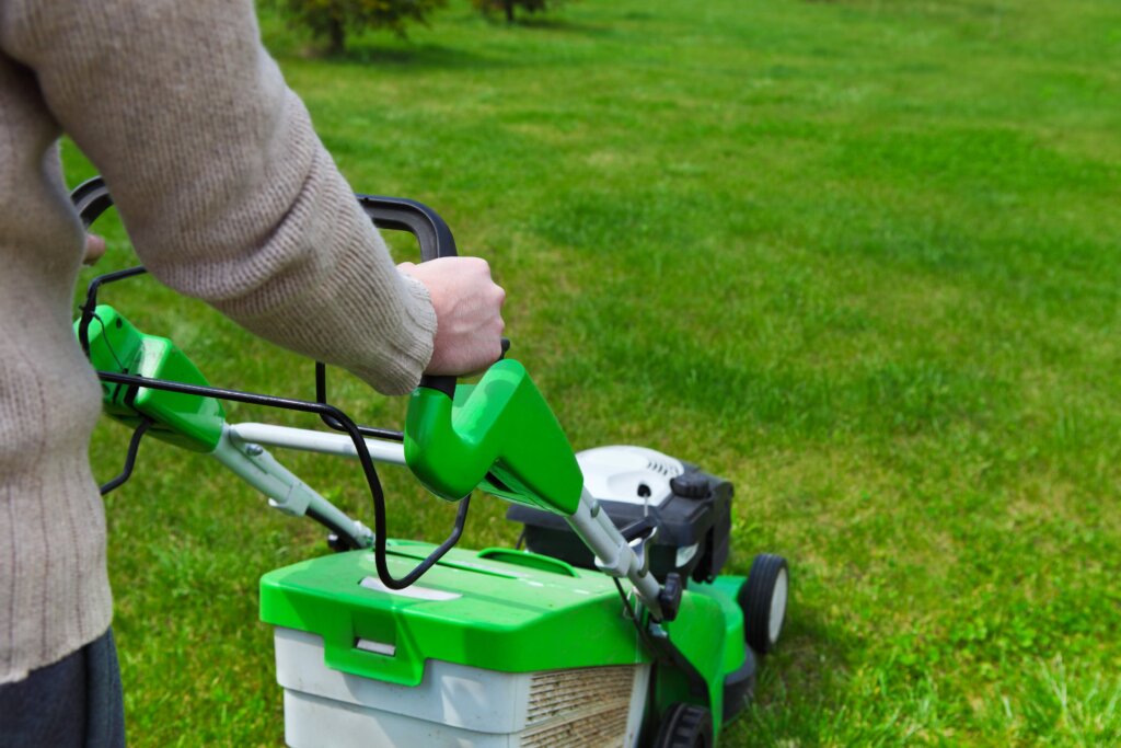 A person wearing a beige sweater is pushing a bright green lawnmower across a lush green lawn, prompting the question: fertilize before or after mowing?