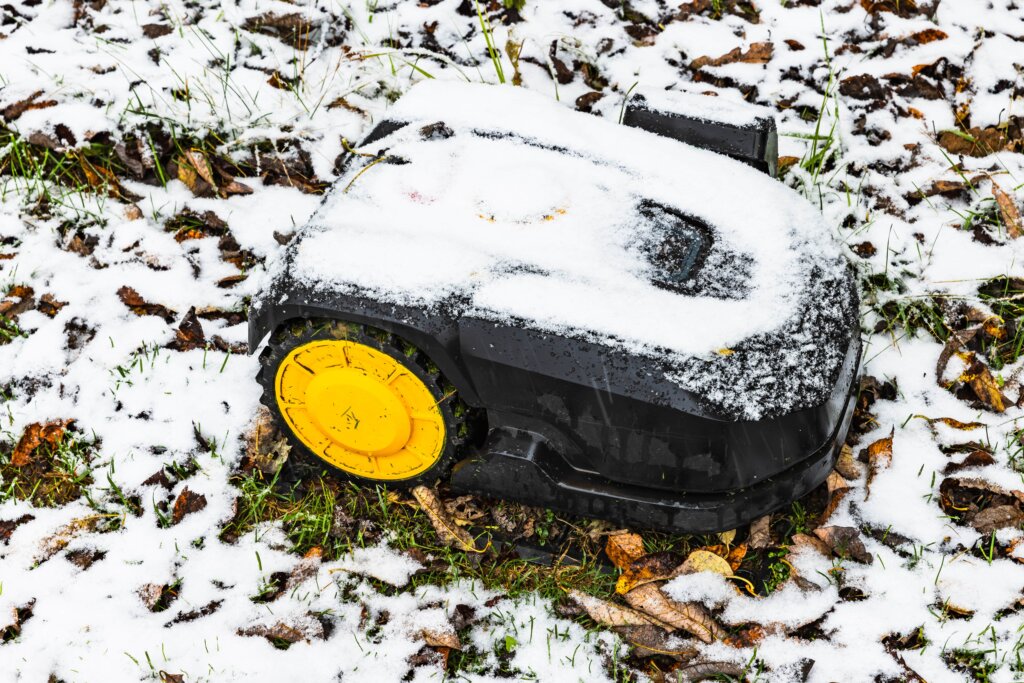 A robotic lawn mower sits on a snow-covered lawn with leaves.
