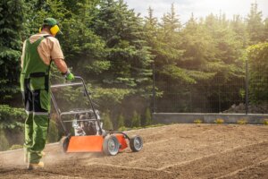 A man wearing overalls and ear protection is operating a core aerator machine on a large, prepared patch of dry, brown soil, demonstrating the process for how to aerate lawn.