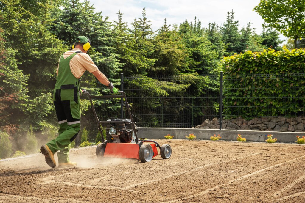 A lawn professional in green work gear is walking behind a powered aerator, kicking up dust on a patch of bare, prepared soil, illustrating the first physical step for how to aerate lawn.
