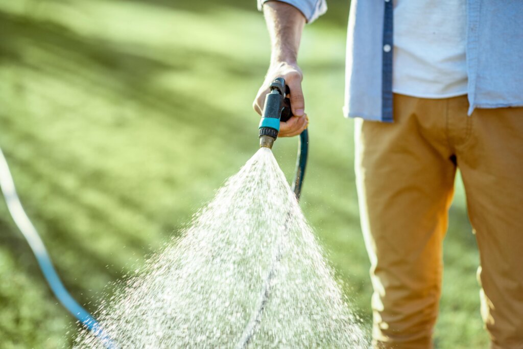 A man holding a running hose and nozzle, spraying a wide cone of water or liquid mixture across a green lawn, demonstrating the application of liquid lawn fertilizer.