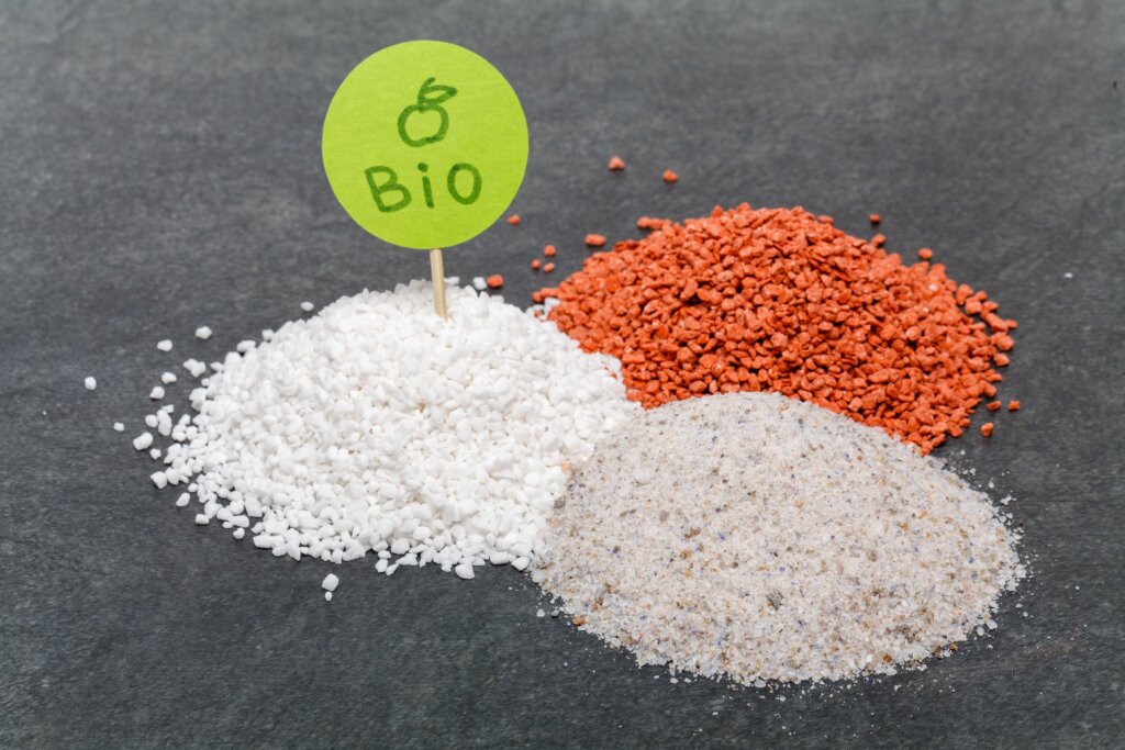 Three separate piles of granular fertilizer—white, red-brown, and gray sand-like—on a dark surface with a green tag reading "BIO," visually representing the components of Nitrogen, Phosphorus, and Potassium Lawn Fertilizer.