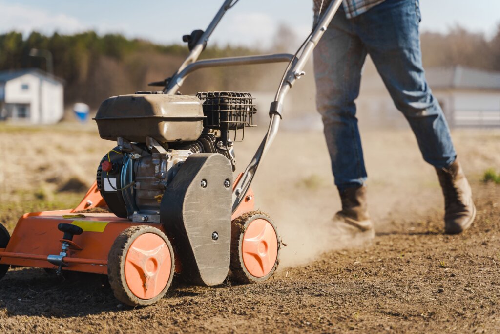 A close-up of a person pushing an orange motorized aerator across a large, dusty field of soil, illustrating the consistent effort needed when deciding when to aerate lawn over several years.