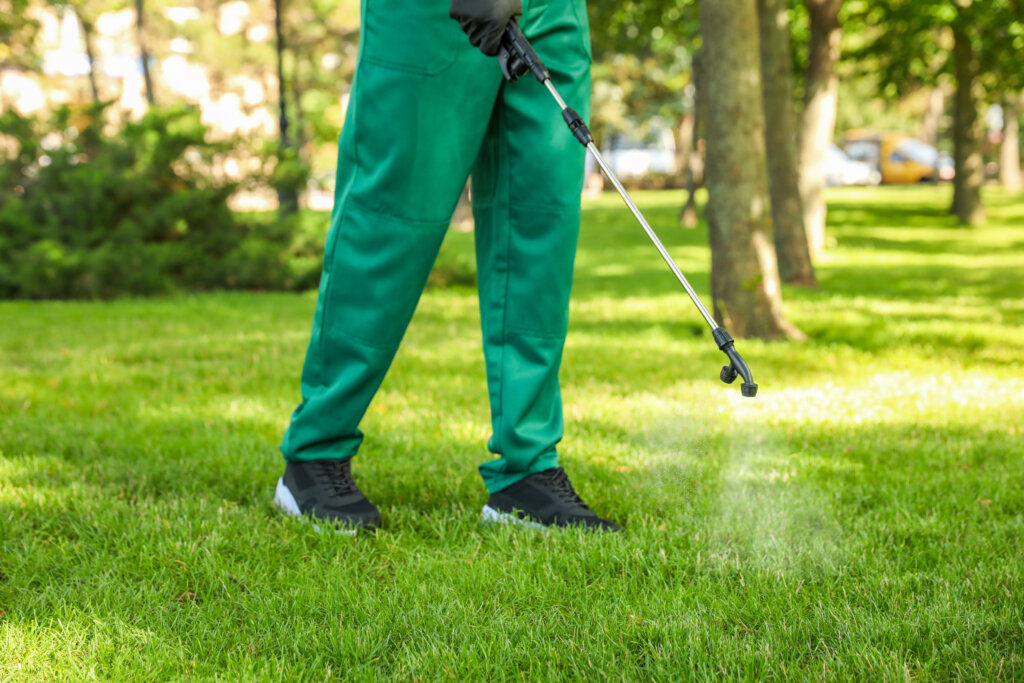 A person in green work pants and black boots stands on a lawn spraying a fine mist from a long wand sprayer onto the grass, with trees and a parking area blurred in the background.
