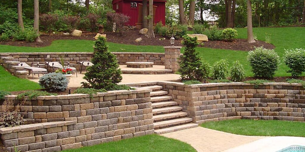 A tiered backyard patio with stone retaining walls and mulch beds backing right up to dense mature woods