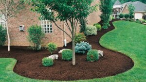 A curved mulched bed wraps around a brick house corner, with a young shade tree, boulders, and shrubs set against a bright green lawn.