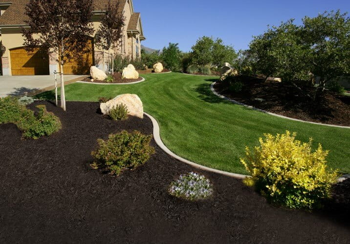 A neatly landscaped front yard showing a strip of bright green grass curving smoothly between dark mulch beds and large decorative rocks next to a house.