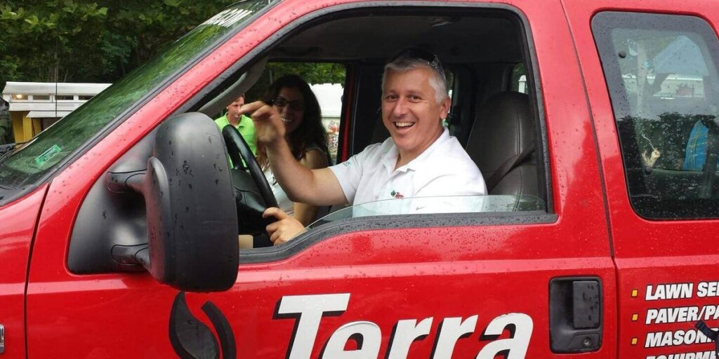 A smiling gray-haired man sits at the wheel of a red Terra work truck, with a woman seated behind him.